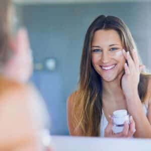 smiling brunette female applying face cream under her eyes in the mirror