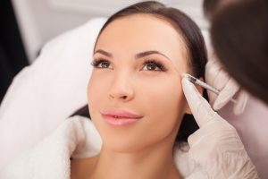 Beautiful young woman is getting botox injection at clinic. She is sitting and smiling. The doctor is holding syringe near her eyebrows carefully