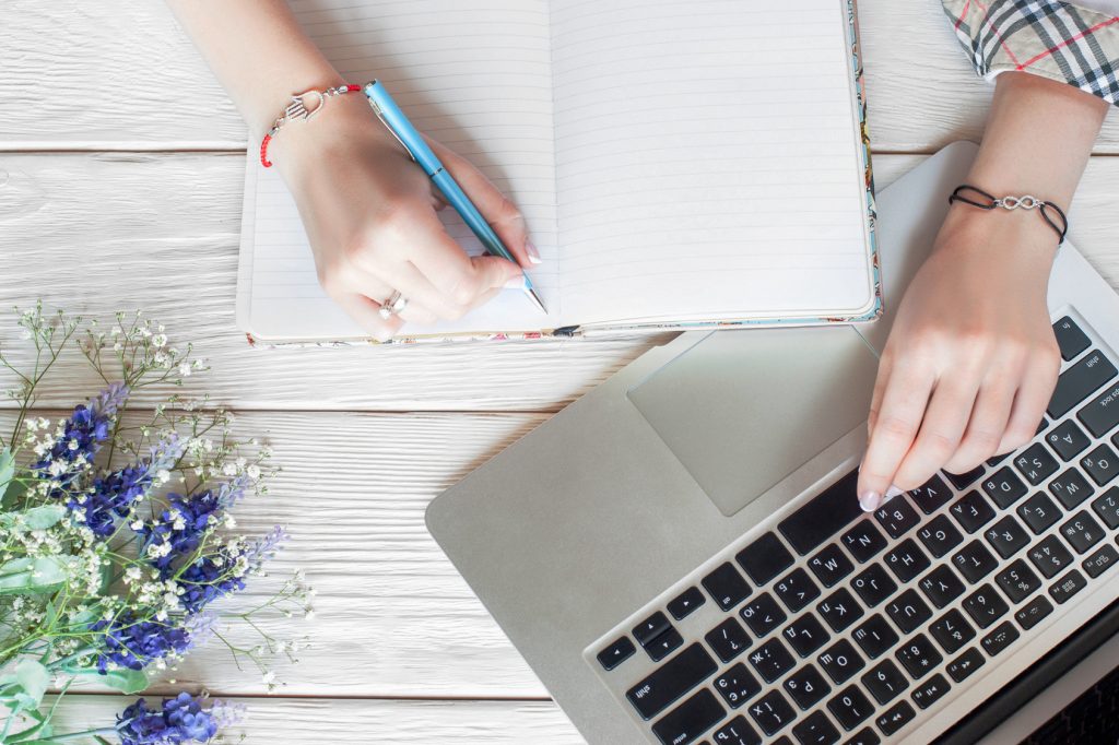 Fashionable business woman working with notebook. Top view on female student hands in cool bracelets, writing in dairy and typing on laptop, flat lay