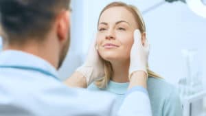 Plastic / cosmetic surgeon examines a beautiful woman's face, touches it with gloved hands, inspecting healed face after facelift surgery with amazing results.