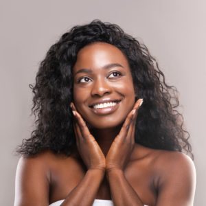 dark skinned female with dark hair smiling against a gray background with her hands up to her face
