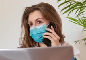 masked female with brunette hair talking on the phone behind a computer